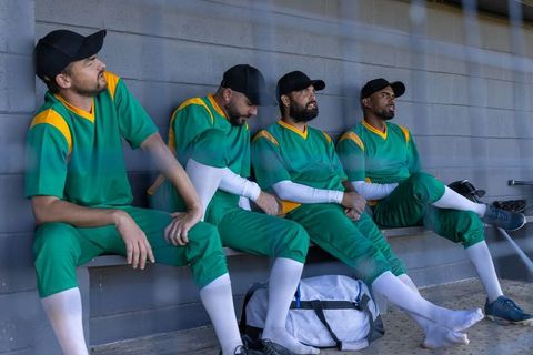Baseball players sitting in dugout wearing green uniforms and black caps