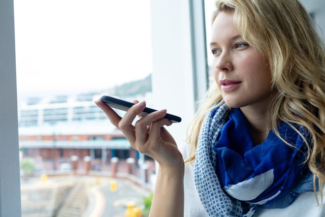 Young businesswoman talking on smartphone near office window, wearing blue scarf. Perfect for illustrating modern communication, professional settings, business environments, and technology use in workplaces.