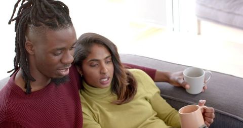 Multicultural couple relaxing on sofa with mugs, sunlit living room, cozy lifestyle