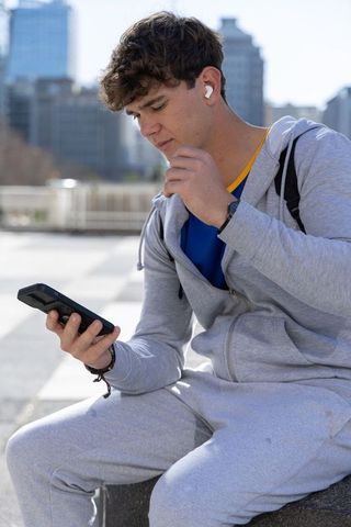 Young man in gray tracksuit adjusting wireless earbud while checking smartphone in city