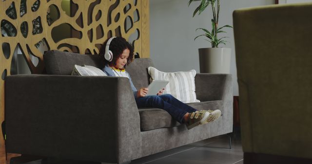 Little boy lounging on plush sofa while using digital tablet and wearing headphones in bright, contemporary living room with stylish decor and plants. This image can be used for home lifestyle, technology in daily life, childhood leisure, and modern living concepts.