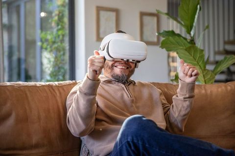 Asian man smiling and interacting with vr headset while relaxing on contemporary living room sofa