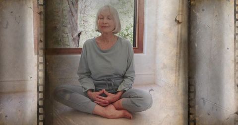 Meditating senior woman practicing mindfulness at home by window in soft grey loungewear