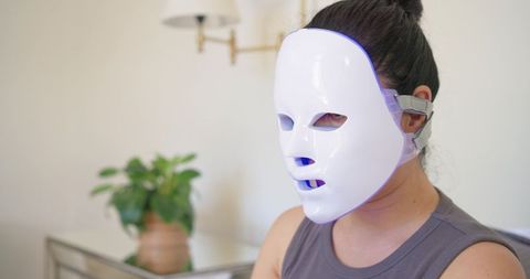 Woman wearing led face mask glowing blue practicing at-home skincare in minimalist interior