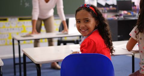 Smiling Schoolgirl Sitting in Classroom with Teacher Near Board