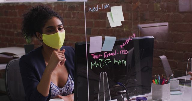 Biracial businesswoman using memo notes sitting in front of computer wearing face mask - Download Free Stock Photos Pikwizard.com