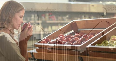 Woman smelling fresh apple while selecting produce from wooden crates in grocery market