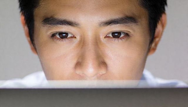 Focused young man studying on laptop late night closeup with screen reflection in eyes