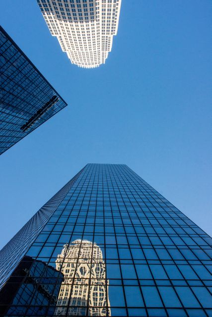 Modern glass skyscrapers reflecting other buildings against a clear blue sky, creating a striking urban landscape. Perfect for websites, articles, and advertisements relating to architecture, urban development, city life, corporate environments, or modernization.