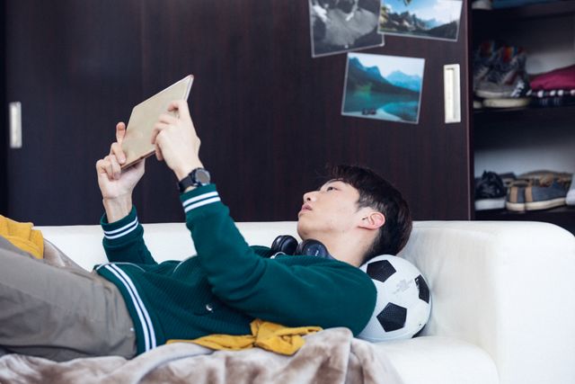Teenage asian boy using digital tablet while lying on couch at home, copy space. Unaltered, people, lifestyle, resting, home and technology concept.