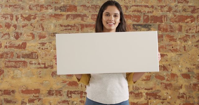 Woman in casual clothing holding blank sign, standing against rustic brick wall. Ideal for advertising, marketing, announcements, or messages. The clean, simple background emphasizes the sign, suitable for various uses, including branding and promotional materials.