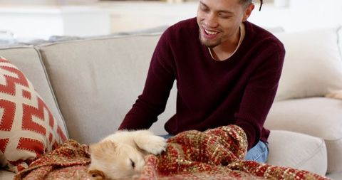 Smiling man petting sleeping puppy on cozy beige sofa with woven blanket