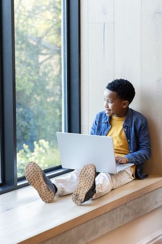 African american boy studying with laptop on window seat, smiling and looking outside