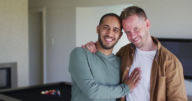 Happy interracial gay couple standing in a modern living room, smiling warmly at camera. This image can be used for promoting LGBTQ diversity, inclusivity, love, and representation. Suitable for use in family life, relationships, and home lifestyle contexts.
