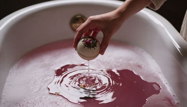 Hand dropping floral bath bomb creating pink water ripples with rose petals