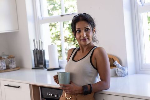 Indian woman leaning on white kitchen counter holding mug and gazing thoughtfully