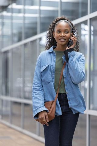 African woman walking and talking on smartphone by glass building wearing denim shirt