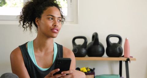 African american woman sitting on floor holding smartphone in home workout space