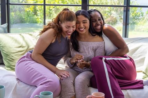 Multicultural friends laughing and hugging in cozy loungewear on sunlit sofa morning chat
