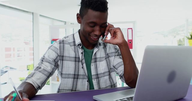 A young African American man is engaged in a phone conversation while working on a laptop, with copy space. His casual attire and the home office setting suggest he might be telecommuting or managing personal business.