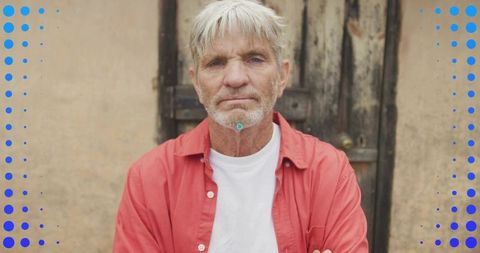 Stoic senior man standing arms crossed wearing red shirt in front of weathered wooden door