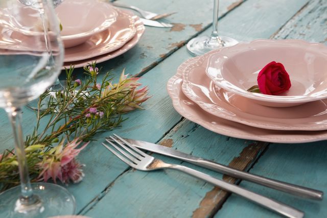 Elegant table setting featuring a red rose placed in a pink bowl on a rustic wooden table. Ideal for use in content related to romantic dinners, special occasions, celebrations, and dining decor inspiration. Perfect for blogs, social media posts, and advertisements focusing on intimate dining experiences and elegant table arrangements.