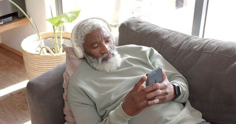 Elderly Man Relaxing on Sofa with Foldable Smartphone and Headphones