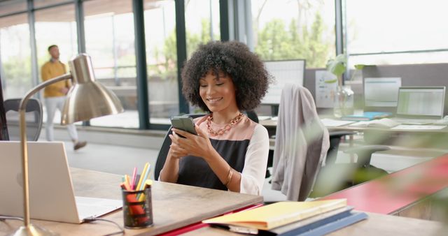 African American professional sitting at desk using smartphone in modern office. She is smiling and appears to be checking messages or social media. The contemporary workspace with open design and desk accessories creates a productive and friendly environment. This can be used to represent modern workplaces, technology in business, professional communication, or inclusive work cultures.