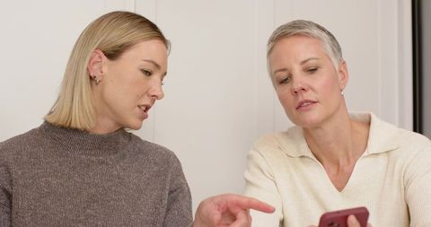 Two women discussing smartphone while pointing, casual coworkers sharing mobile screen