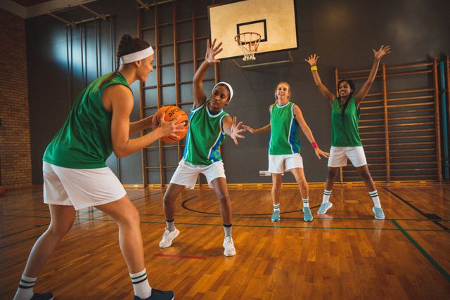 Diverse group of female athletes practicing basketball in an indoor court. One player is preparing to shoot while others are defending. Ideal for use in sports training materials, teamwork promotions, athletic apparel advertisements, and articles on women's sports.