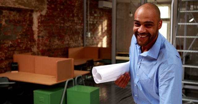 Architect standing in an urban office holding blueprints and smiling to camera. Brick walls and modern furniture are visible in the background, suggesting an industrial design setting. Ideal for use in office, architecture, and professional business contexts, emphasizing modernity and creativity.