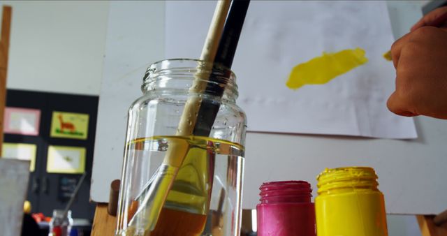 An artistic workspace featuring brushes in jars, vibrant yellow paint being applied to paper. Ideal for themes of creativity, education, art classes, and hobbies. Suitable for promoting art supplies, educational materials, and creative workshops.