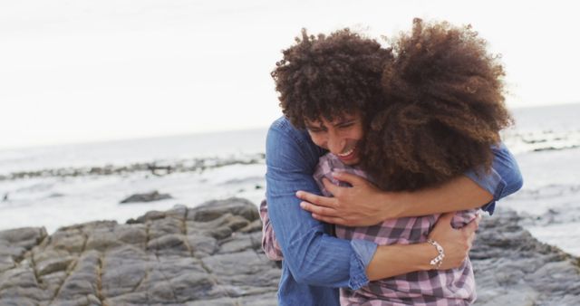 This photo depicts a joyous African American couple hugging on a rocky beach, with the ocean in the background. Their happy expressions and close embrace convey emotions of love and connection. This can be used in content related to romance, relationships, travel, vacations, and outdoor activities.