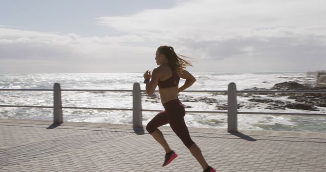 Woman dressed in athletic wear running along a seaside promenade on a bright day. The ocean waves and open sky create a picturesque background, ideal for promoting fitness, active lifestyles, and healthy living. Suitable for use in health and wellness marketing, fitness app promotions, exercise blogs, and outdoor activity advertisements.