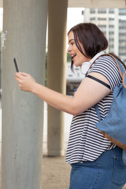 Curvy Caucasian woman out and about in the city streets during the day, laughing and using her smartphone wearing a backpack and headphones, with a modern building in the background