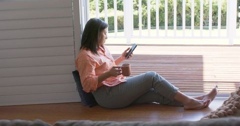 Asian mature woman relaxing at home on doorway with coffee and smartphone on sunlit deck