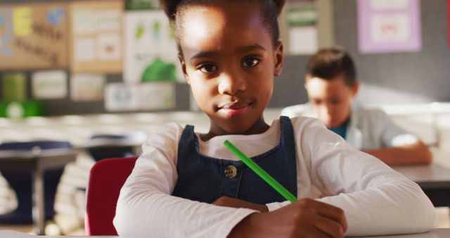 This image captures a young student attentively writing in a classroom setting. The child, sitting on a red chair, is dressed in a white shirt and denim overalls, holding a green pencil. The background features another student focusing on their work. Ideal for educational materials, school or learning concept advertisements, websites, or blogs focused on childhood education, diversity, and school activities.