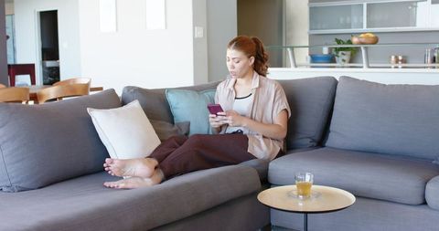 Young woman relaxing and using smartphone on modern grey sofa in minimalist living room