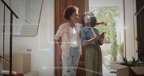 Young women moving into new home, smiling and embracing at doorway with boxes and smartphone