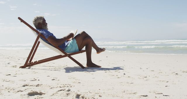 Senior man is relaxing on a beautiful sandy beach while reading a book. He is sitting in a wooden lounge chair facing the ocean with gentle waves in the background. Ideal for materials promoting beach vacations, leisure activities, older adults finding relaxation, or book reading during summertime.