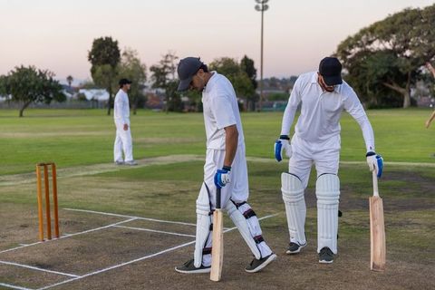 Indian and asian teammates walking across worn cricket pitch at dusk carrying bats