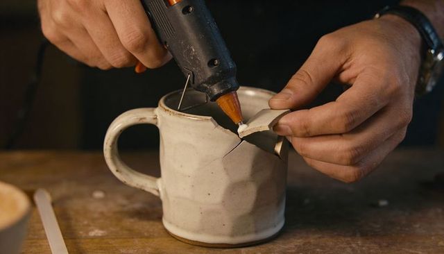 Repairing cracked ceramic mug with hot glue gun close-up of hands mending pottery