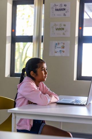 Young girl in pink hoodie concentrating on laptop in bright classroom with windows