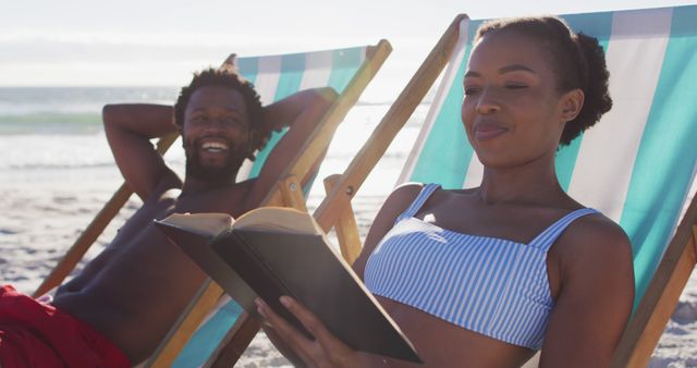 African american couple sunbathing and reading book while sitting on deck chairs at the beach. travel romantic vacation lifestyle concept