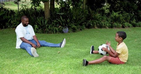 African american father and son sitting on lawn playing soccer, backyard family bonding