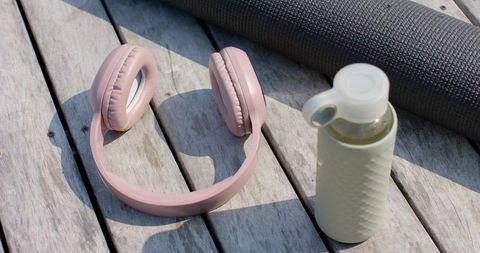 Pastel pink over-ear headphones with water bottle and yoga mat on sunlit wooden deck