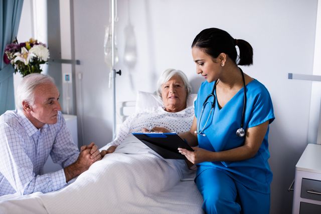 Female doctor in blue scrubs discussing a medical report with a senior couple in a hospital ward. The elderly woman is lying in a hospital bed while the elderly man is sitting beside her. This image can be used for healthcare, medical consultation, elderly care, and hospital-related content.