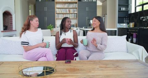 African american and multiracial women chatting on sofa with mugs in open-plan kitchen