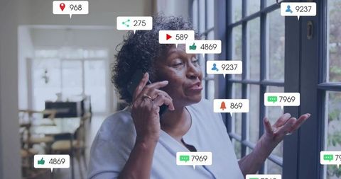 Senior woman talking on smartphone by sunlit glass doors with social notification icons