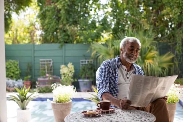 Senior man enjoying a quiet morning in his garden, reading a newspaper while sitting at a table. Ideal for use in articles or advertisements related to retirement, senior lifestyle, relaxation, and outdoor activities.